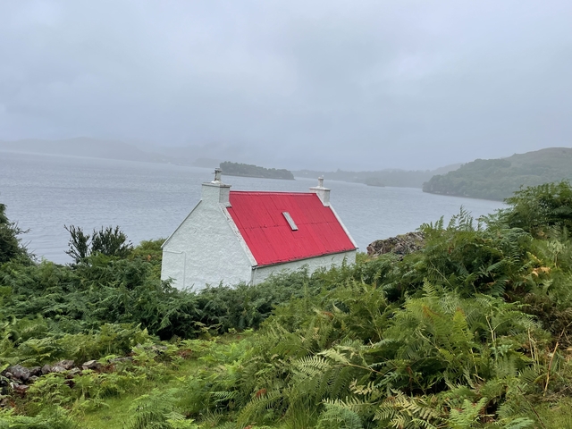       A small white house with a red roof surrounded by greenery, overlooking a body of water.
  