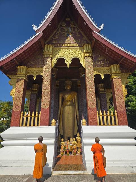 Monks in traditional attire stand before a large golden Buddha.