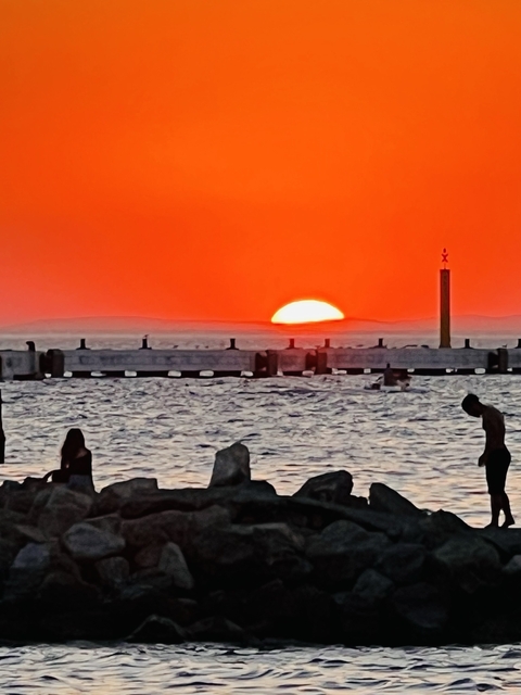       A vivid orange sunset over the sea with people silhouetted.
  