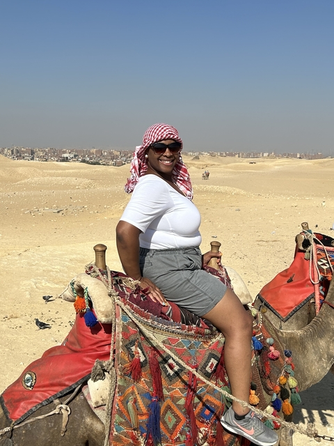 Person on camel in a desert with a city in the background.