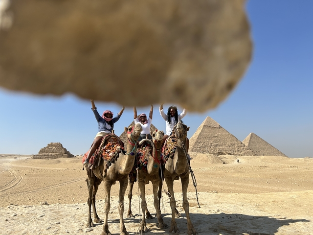 People on camels with pyramids and a rock in the foreground.