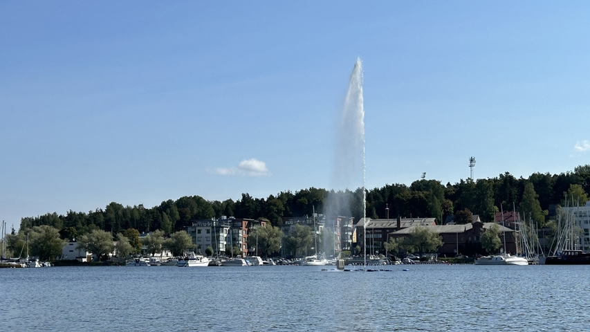       Fountain spraying water in a lake with surrounding buildings.
  
