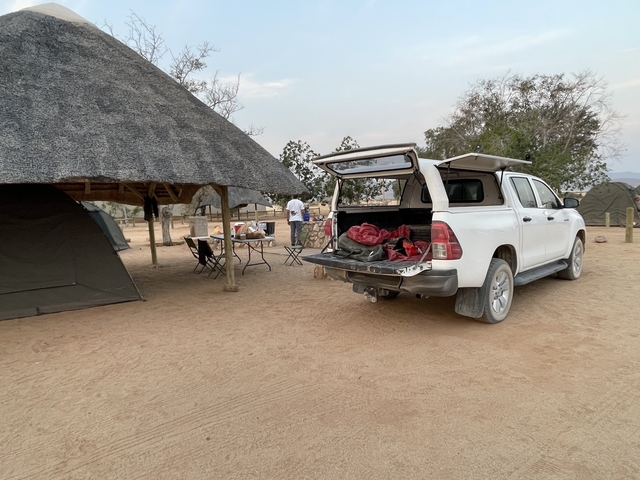 Campsite with car parked next to a tent and thatched roof shelter.