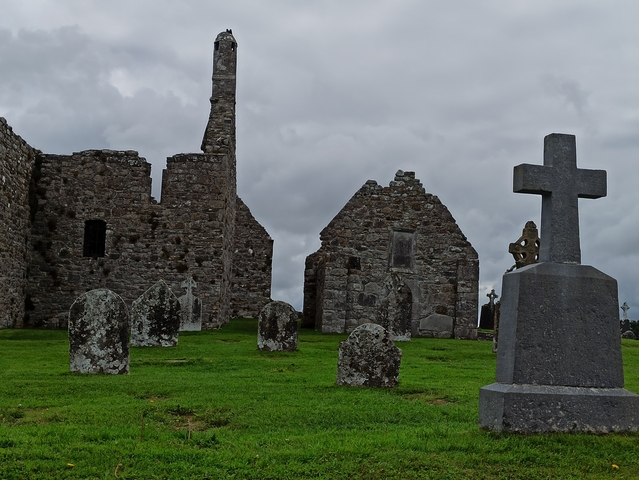 Ancient stone ruins with gravestones in a grassy field under a cloudy sky.