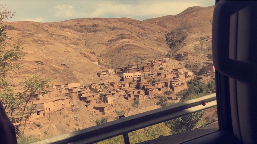       View of a village built on a hillside with brown buildings and a metal guardrail in the foreground.
  