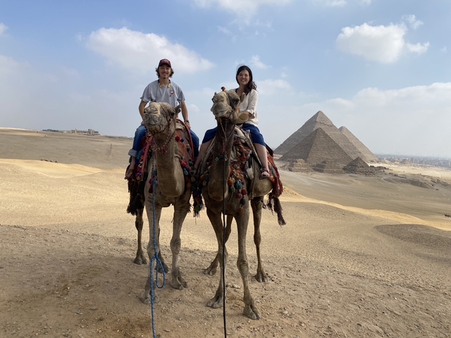 Couple riding camels in front of pyramids.