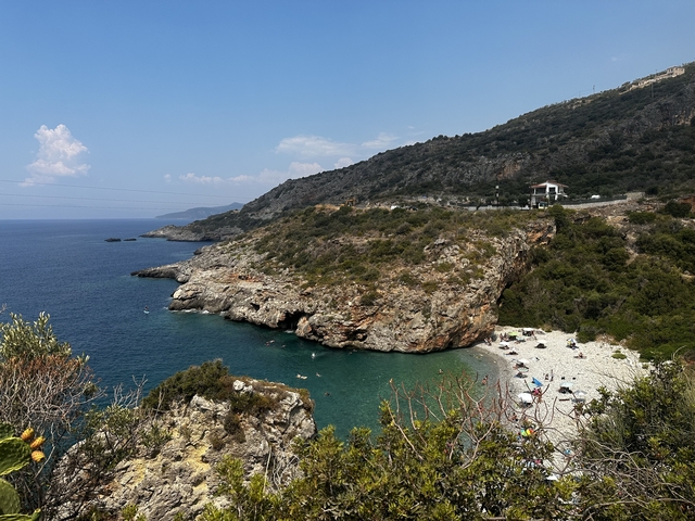       Pebble beach with people sunbathing, surrounded by cliffs and blue sea.
  