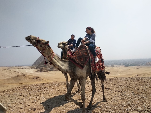 People riding camels in the desert with pyramids in the background.