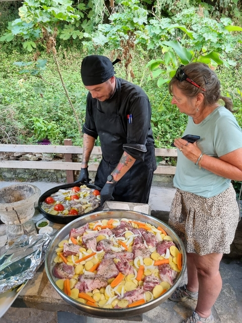       Chef preparing a dish outdoors with a woman observing.
  