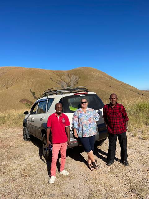       A group of people posing with a car in a mountainous area.
  