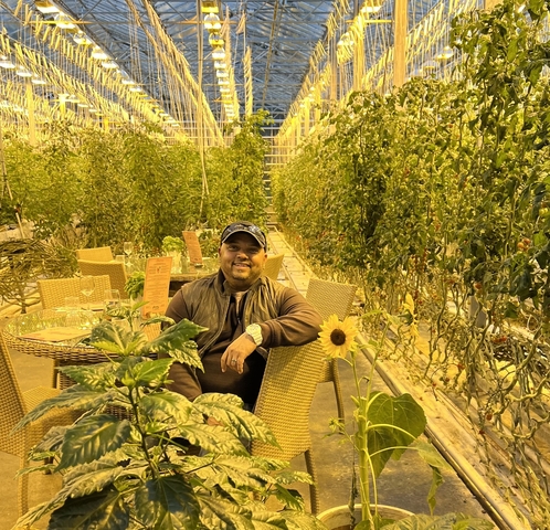       Man sitting among plants inside a greenhouse.
  