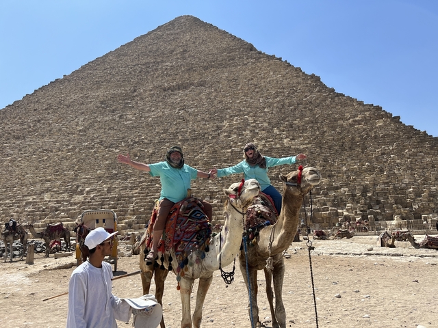 Two people riding camels in front of a pyramid.