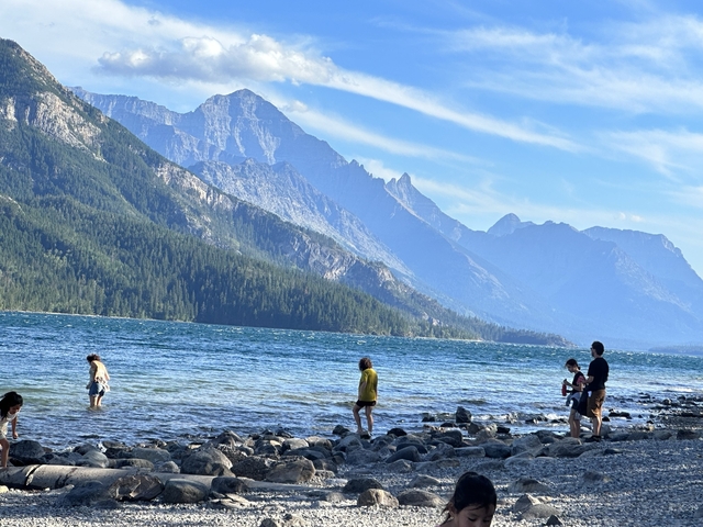 People walking along a rocky lake shore with mountains in the background.