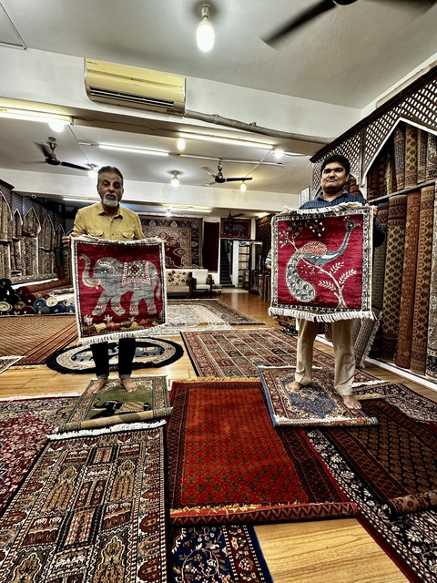 Two men displaying decorative rugs inside a shop.