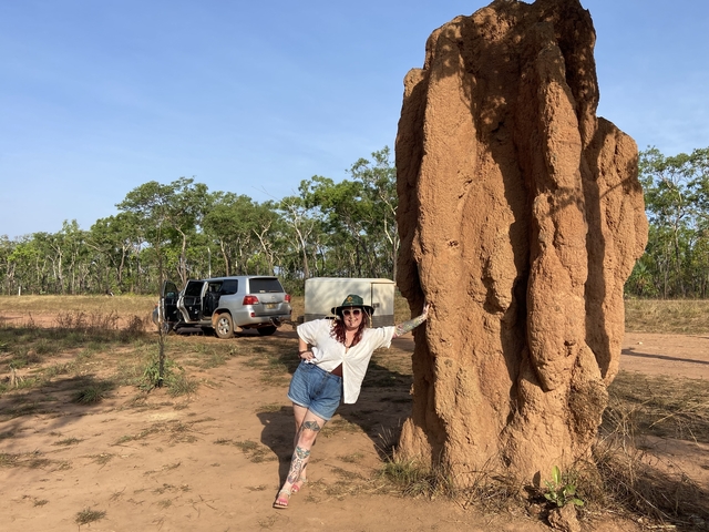 Person posing next to a large termite mound in a savannah landscape.