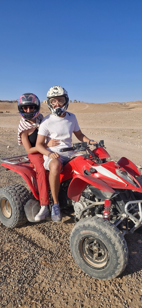       Two people on a quad bike with helmets.
  