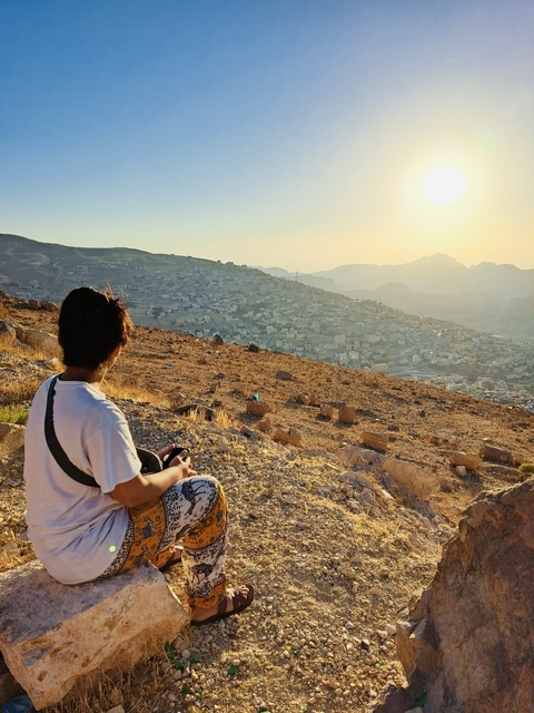 Person seated, looking over a city nestled in hilly terrain at sunset.
