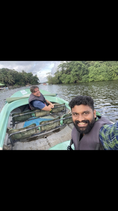 Two smiling men in a boat on a river with surrounding trees.