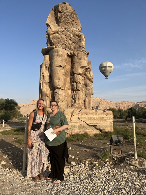       Two people posing with a statue and a hot air balloon in the sky.
  