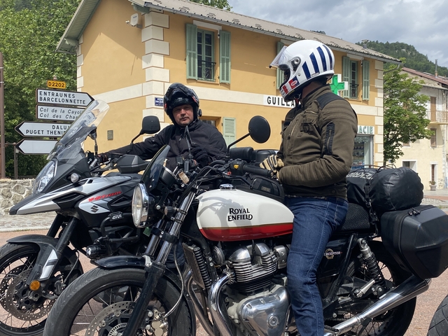       Two individuals on motorcycles in front of a village sign.
  