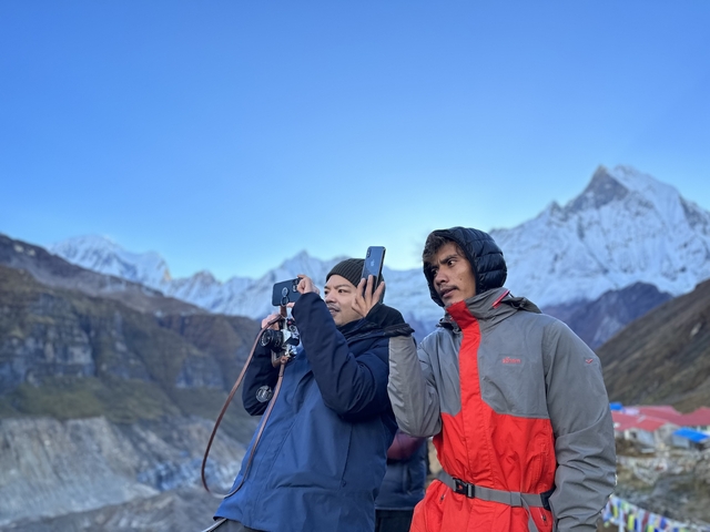       Two people capturing a view of snow-capped mountains.
  