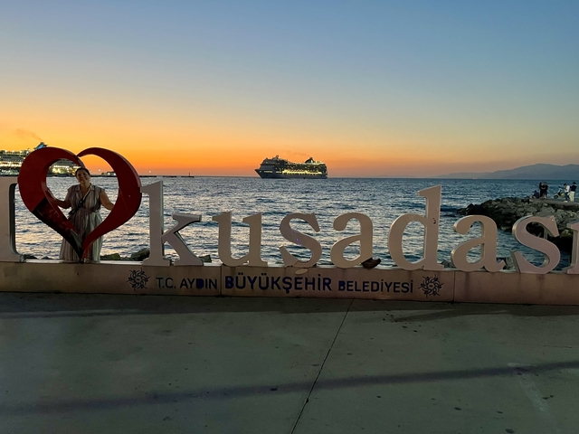       Woman posing beside a sign with a cruise ship in the background during sunset.
  