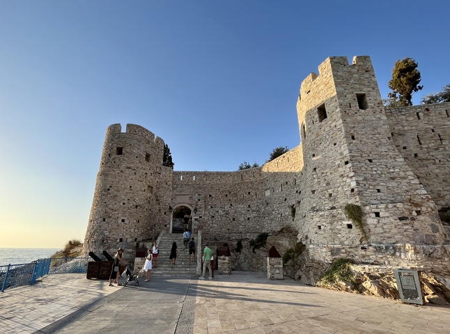      Stone castle by the sea with people around.
  