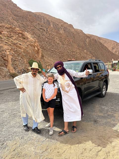       Group of people posing with a car in a desert landscape.
  
