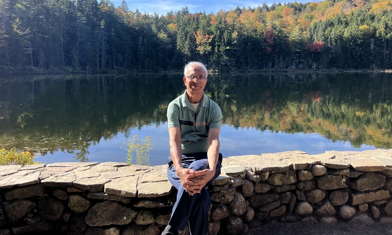 Man sitting on a stone wall by a lake with trees in the background.