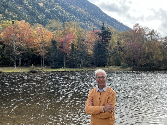 Man standing by a lake with vibrant autumn trees.