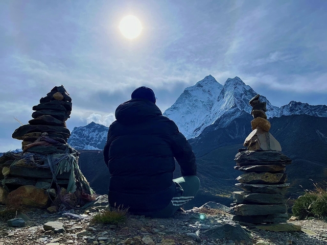 Person sitting between stone cairns with mountain backdrop.