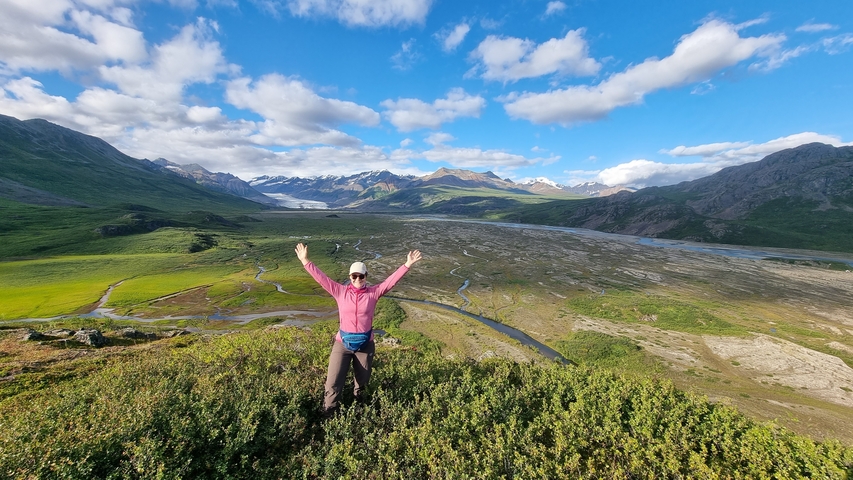       Person posing in a vast landscape with mountains and river.
  