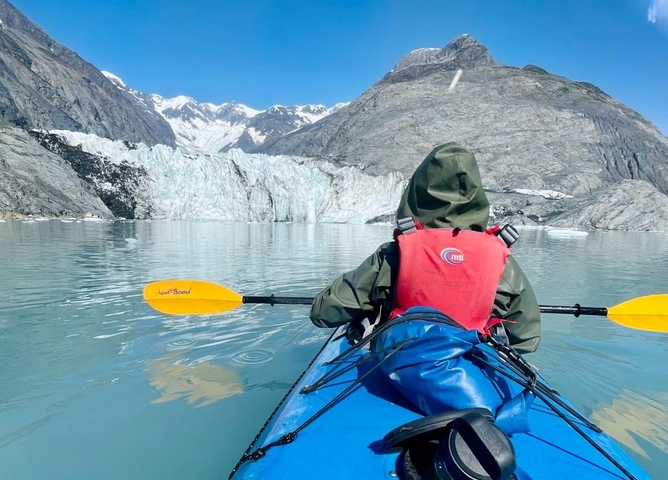       Person kayaking towards a glacier in a scenic mountain landscape.
  