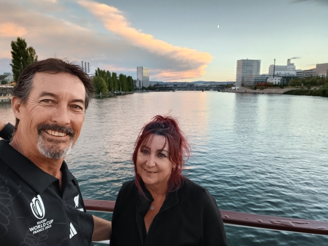      Smiling couple posing by a river with cityscape.
  