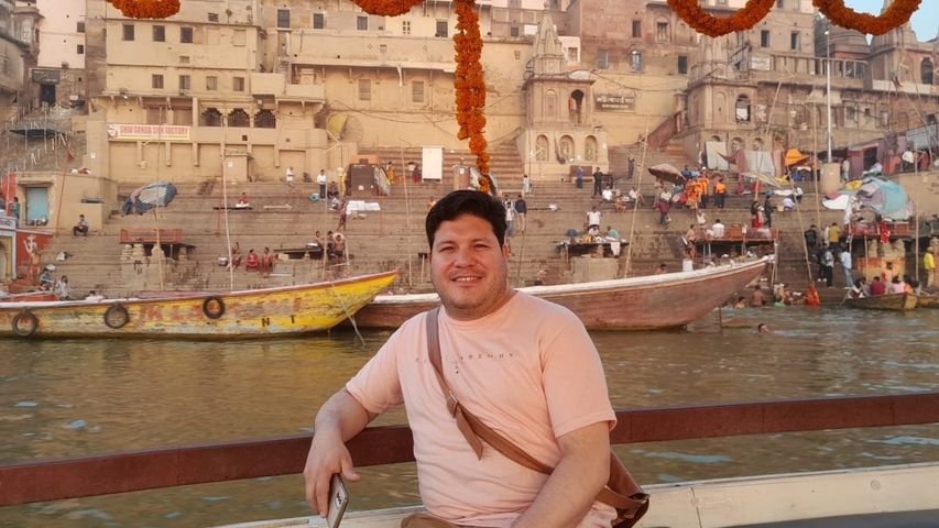 Man sitting on a boat in front of ghats with boats and people.