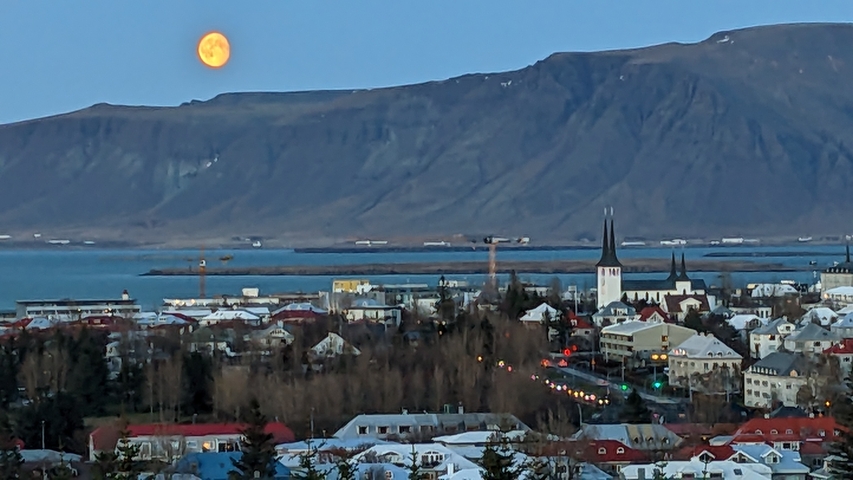       Panoramic view of a city with mountains and a large moon.
  