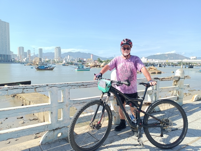       Bicyclist posing by a river with city buildings in the background.
  
