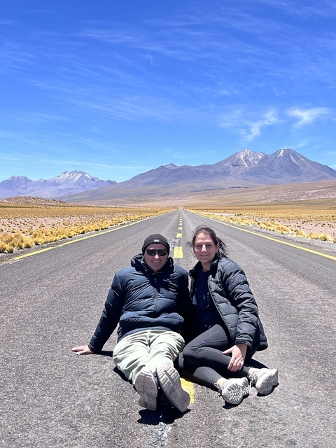       Two people sitting on a road with mountains in the background.
  
