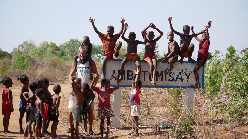 Group of children and a man posing on a sign in a rural area.