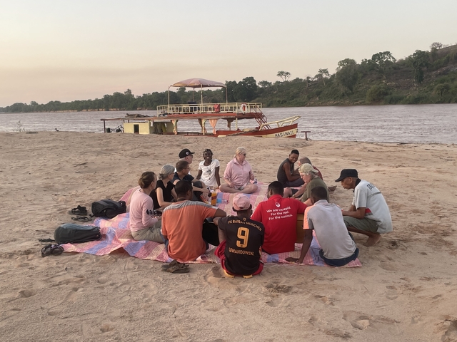 Group of people having a picnic by a riverside with a boat nearby.