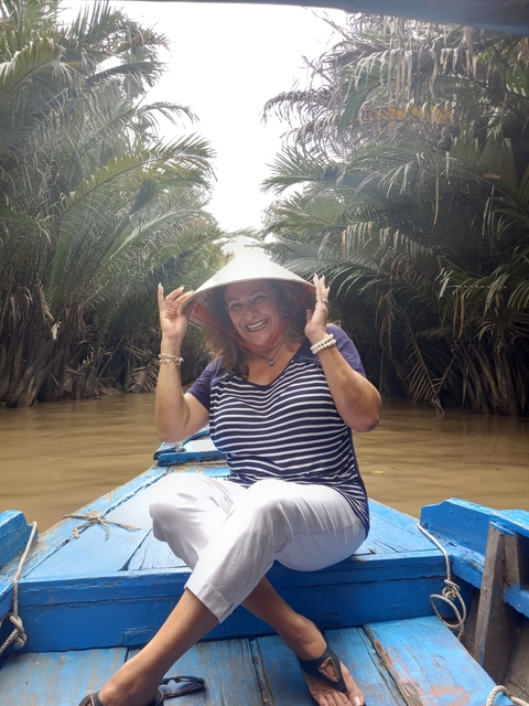 Woman wearing a traditional hat on a boat in a river surrounded by palm trees.