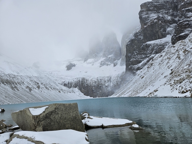 Turquoise glacial lake surrounded by snowy mountains and mist.
