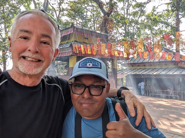       Two people posing in front of a small shrine with prayer flags.
  