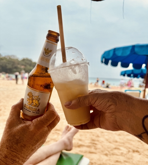 Two people toasting with a beer and a cocktail on a beach.