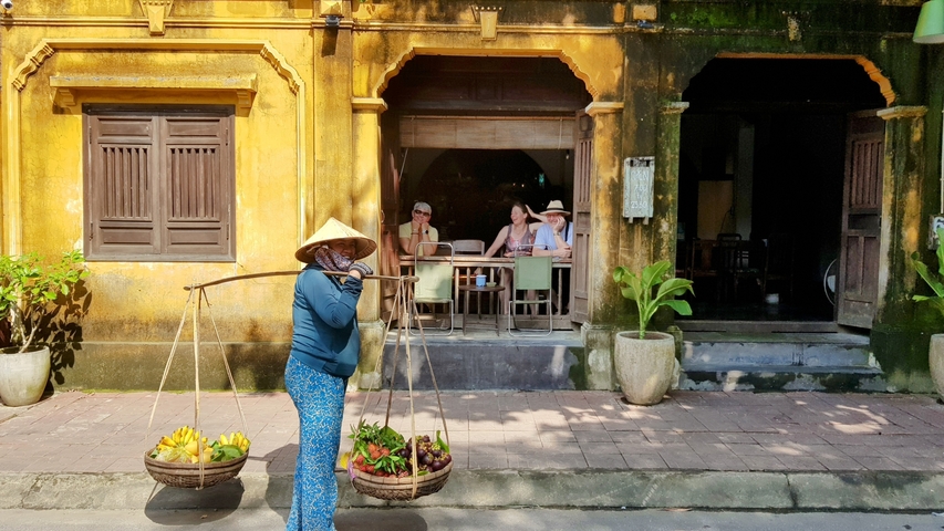 Woman carrying baskets of produce in front of a yellow building