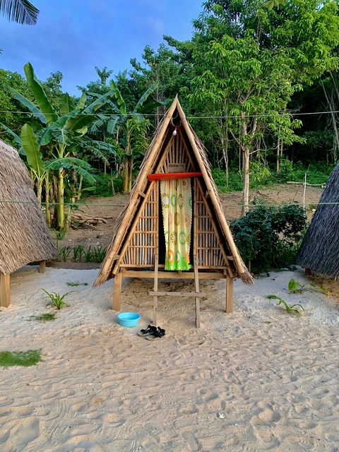 A triangular bamboo hut on a sandy path surrounded by lush greenery.