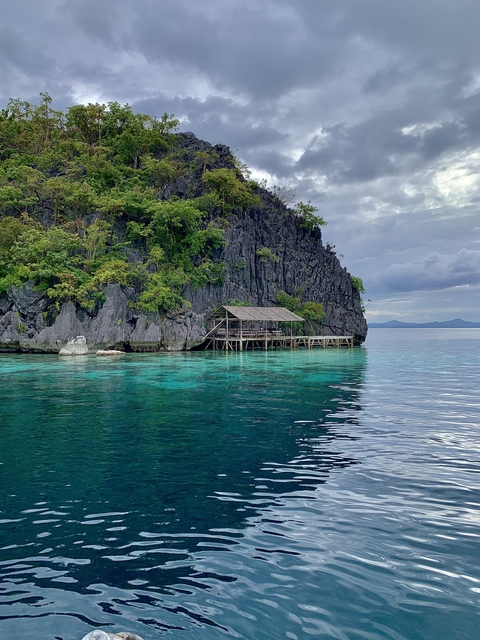 A stilt house over clear blue water, with a rocky cliff covered in vegetation in the background.
