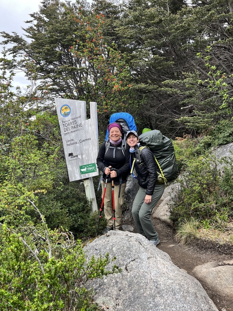 Two people posing by a sign with hiking gear, in Torres del Paine National Park.