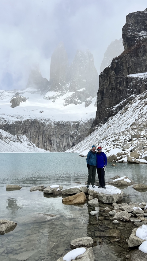 Two people standing near a glacial lake surrounded by snow-capped mountains.