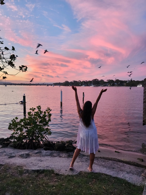       Woman with arms raised embracing a picturesque pink sunset by the water.
  